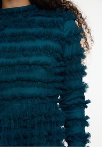 Teal mekko, joka on valmistettu kerroksellisesta satiinista ja röyhelöistä, pitkähihainen ja tekstuuripinta tuo syvyyttä ja ulottuvuutta.
