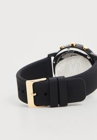 Bracelet de montre en caoutchouc noir avec une finition texturée, fermoir en métal doré, et boîtier rond en métal noir avec des accents dorés. Cadran blanc détaillé à l'arrière.