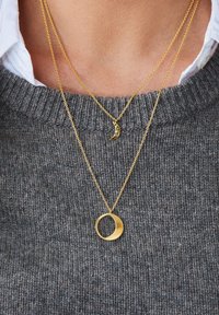 Vergoldete Halsketten mit einem kleinen Halbmond und einem runden Anhänger, beide an feinen Ketten aufgehängt, vor einem grauen Pullover.