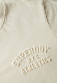 Sudadera color crema con un logo texturizado en relieve que dice "SUPERDRY N.Y.C. ATHLETICS" en la parte frontal. Acabado de tela suave.