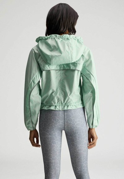 Kvinde iført en mintgrøn hættetrøje og grå leggings med blomsterprint, stående med ryggen mod, foran en ensfarvet baggrund.