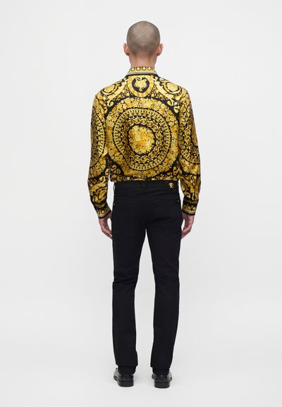 Homme à la tête rasée portant une chemise noire et or à manches longues à motifs, un pantalon noir et des chaussures noires, debout de dos.