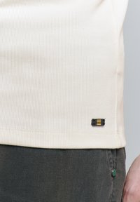 Cr�ème geribbelde shirt met een zwart label met gele accenten aan de onderkant, gecombineerd met donkergrijze stof met textuur.