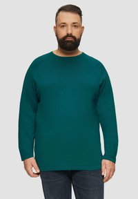 Pull long à manches longues teal avec un col rond, texture côtelée et ourlet droit. Porté avec un jean bleu foncé, présentant des coutures discrètes.