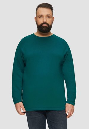 Pull long à manches longues teal avec un col rond, texture côtelée et ourlet droit. Porté avec un jean bleu foncé, présentant des coutures discrètes.