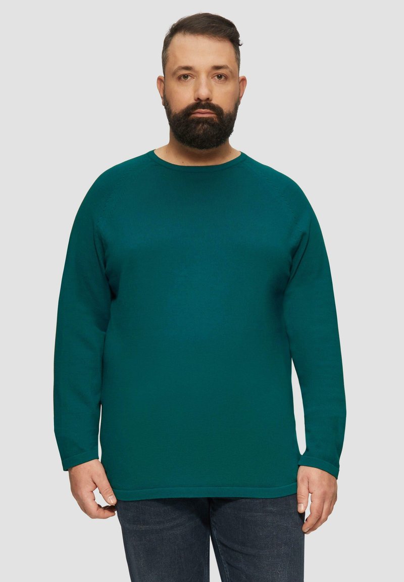 Pull long à manches longues teal avec un col rond, texture côtelée et ourlet droit. Porté avec un jean bleu foncé, présentant des coutures discrètes.