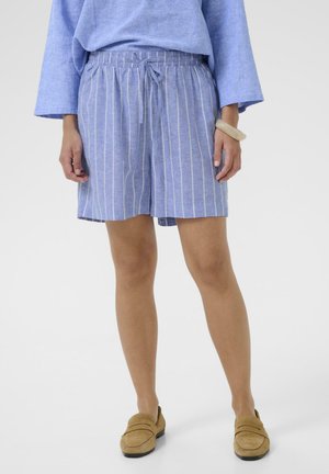 Person iført blå langærmet bluse med elastisk talje, blå og hvide stribede shorts og beige loafers, stående med hænderne afslappet ned langs siderne.