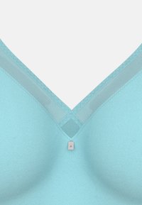 Světle modrá podprsenka s V-čkovým designem a síťovými detaily, se stříbrnou mažou uprostřed. Hladký povrch s bezproblémovým provedením.