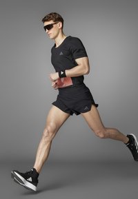 Zwarte sportshirt en shorts, zonnebril en hardloopschoenen met witte accenten. Hardloper in beweging, met een rood startnummer om de taille.