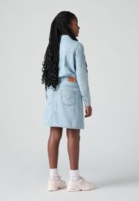 Jovem mulher com cabelo longo trançado, usando uma jaqueta de denim azul claro e saia, meias brancas e tênis brancos, voltada para o lado.