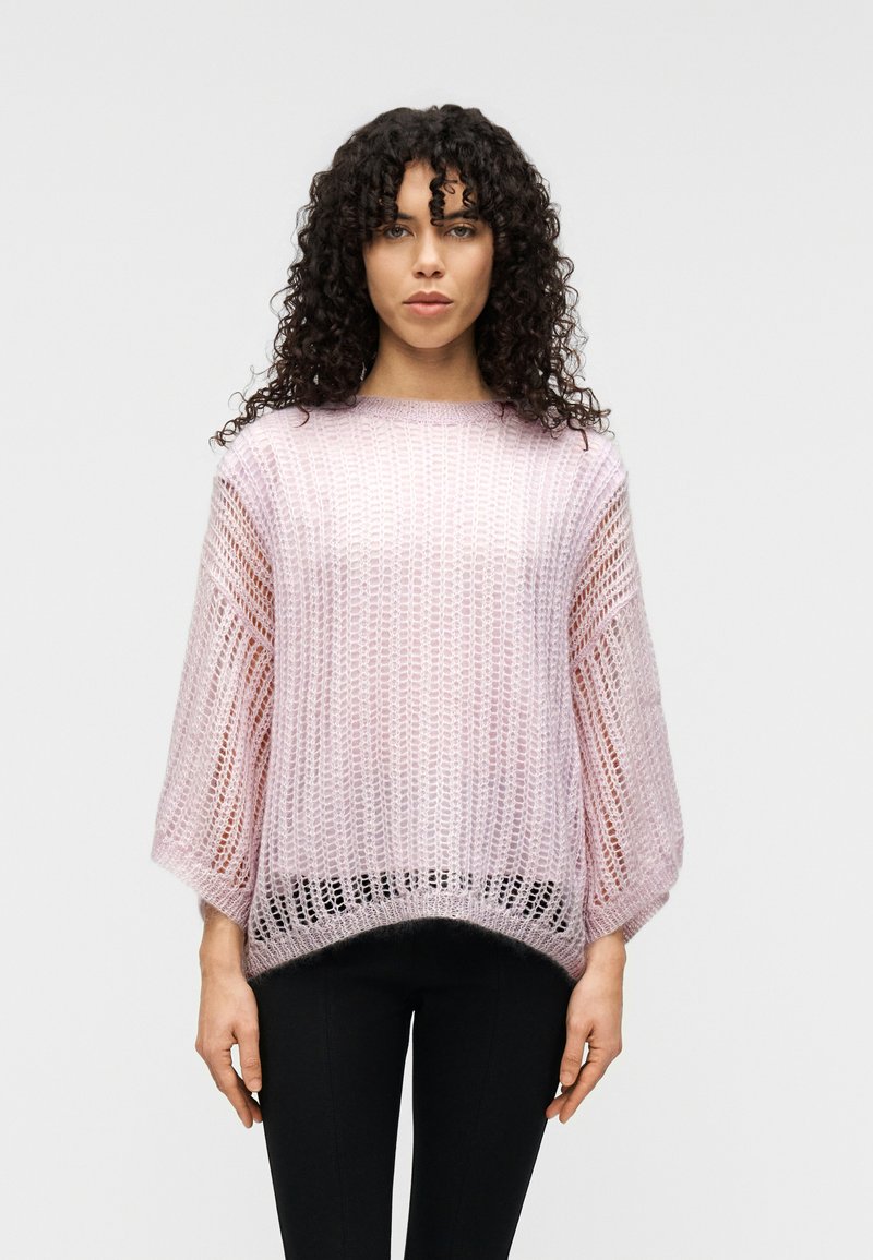 Ανοιχτό ροζ oversized πλεκτό πουλόβερ με φαρδιές μανσέτες, υφή ανοιχτού πλέγματος και στρογγυλεμένο κάτω μέρος, σε συνδυασμό με στενά μαύρα παντελόνια.