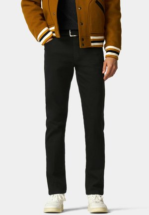 Schwarze gerade geschnittene Hosen mit glatter Textur, kombiniert mit einer braunen Collegejacke und weißen Sneakers. Die Jacke hat gestreifte Bündchen.
