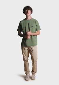 Green cotton t-shirt with a chest pocket and elephant graphic, paired with beige pants and light sneakers, standing against a white background.