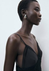Camisole noire avec bordures en dentelle, à fines bretelles. Le mannequin porte une boucle d'oreille métallique brillante. Texture de peau lisse, cheveux courts. Arrière-plan neutre.