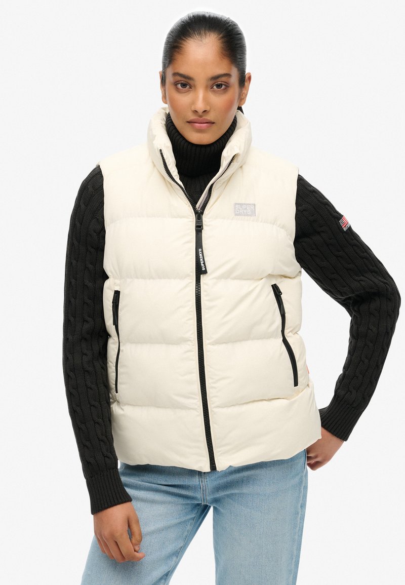 Gilet bianco imbottito con zip nera, tasche laterali e collo alto; abbinato a un maglione nero a trecce e jeans blu.