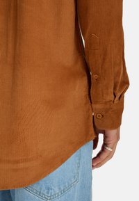 Bruine corduroy overhemd met verticale ribstructuur, met knoopmanchetten en een ronde zoom, gedragen over lichtblauwe denim broek.