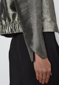 Metalen zilveren jas met een gestructureerde afwerking, voorzien van een gegolfde zoom en gespleten mouwen, gecombineerd met zwarte broek.