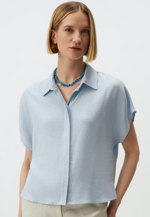 Camicia azzurra a maniche corte con bottoni e colletto, caratterizzata da una vestibilità rilassata e una texture liscia. Abbinata a una collana colorata di perline.