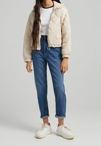 Světle béžová prošívaná bunda s kapucí, elastickými manžetami a zipovým zapínáním vpředu, v kombinaci s modrými džíny a bílými teniskami.