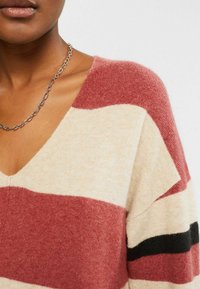 Gestreifter Pullover in Rost-, Beige- und Schwarztönen. V-Ausschnitt mit Strickstruktur. Verfügt über weite Ärmel und eine lockere Passform.