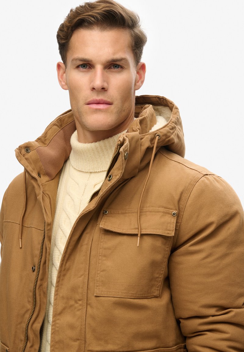 Bruine geïsoleerde jas met een capuchon, voorzien van een rits aan de voorkant, twee borstzakken en een fleece voering. Draag deze over een crèmekleurig kabeltrui.