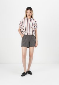 Kurzärmliges, knopfbares Hemd mit roten und blauen Streifen, kombiniert mit grauen Shorts. Das Modell steht mit den Händen in den Taschen und trägt schwarze Schuhe.
