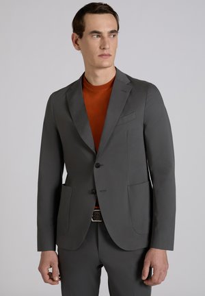 Man in grijs colbert en broek met roestrode overhemd, kijkt naar zijn rechterkant tegen een effen grijze achtergrond.