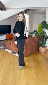 Fekete, testhez simuló garbó, fekete nadrág és szürke papucs, egy világos szobában állva, ahol egy barna kordbársony kanapé és szobanövények találhatóak.