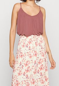 Femme portant un top sans manches mauve et une jupe blanche taille haute avec des motifs abstraits roses et violets, nouée à la taille.