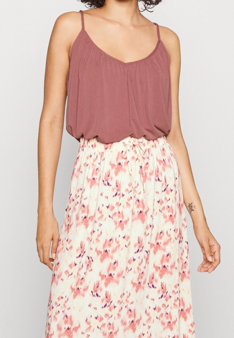 Femme portant un top sans manches mauve et une jupe blanche taille haute avec des motifs abstraits roses et violets, nouée à la taille.