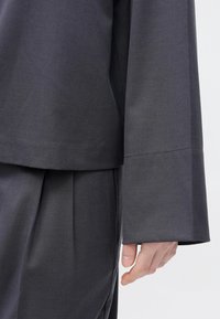 Tmavomodrá, voľne strihaná blúzka so širokými rukávmi a rovný lem, kombinovaná so zladenými plisovanými nohavicami. Hladká textúra a minimalistický dizajn.