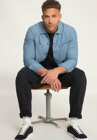 Lichtblauwe jeanshemd met twee borstzakken, gecombineerd met zwarte broek. Zwarte sneakers met witte zolen en witte sokken. Zittend op een kruk.