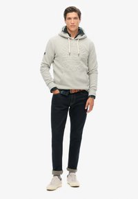 Sudadera gris con capucha y logo en relieve, superpuesta sobre una camisa de cuadros verde, combinada con jeans oscuros y zapatillas crema.