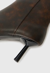 Chaussure à talon haut en cuir marron avec un talon noir brillant. La tige présente une texture lisse avec un léger marbrage et des détails de couture.