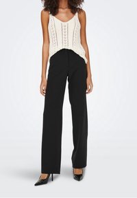 Débardeur crème en dentelle avec col en V et motif tricoté vertical, associé à un pantalon noir à taille haute et jambes larges, et des escarpins noirs pointus.