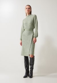 Robe tricotée en vert clair, avec un col montant, des manches longues et une fente sur le côté. Associée à des bottes en cuir noir hautes. Texture lisse.