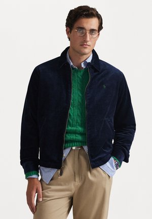 Navy corduroy jacket with a collared neck, layered over a green cable-knit sweater. Light blue and white striped shirt underneath.