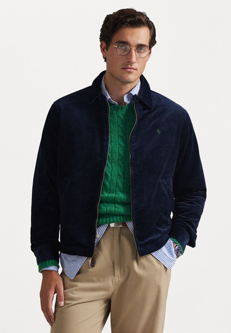 Casaco de veludo cotelê azul-marinho com gola, sobre um sweater verde em ponto cordões. Camisa listrada em azul claro e branco por baixo.