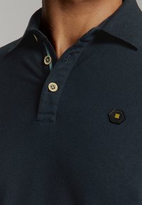 Marineblauwe poloshirt van textuurstof met een drie-knops sluiting. Bevat een zwart hexagonaal logo met gele accenten en een contrasterende binnenkraag.