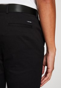 Schwarze Hose mit strukturierter Oberfläche, mit einem dezenten Calvin Klein-Label, einer Seitentasche und einem schwarzen Gürtel.