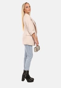 Beige blazer met opgerolde mouwen over lichtblauwe skinny jeans, gecombineerd met zwarte enkellaarzen en een beige handtas met gouden accenten.