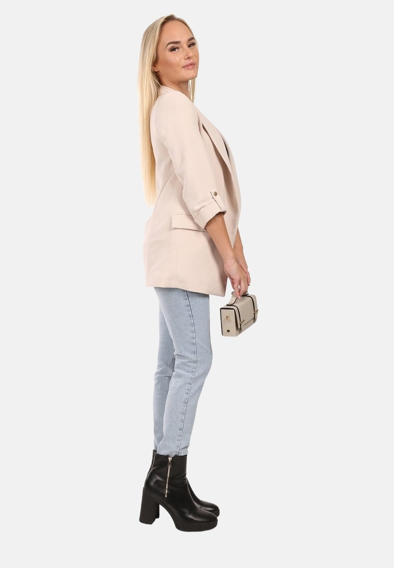 Beige blazer met opgerolde mouwen over lichtblauwe skinny jeans, gecombineerd met zwarte enkellaarzen en een beige handtas met gouden accenten.