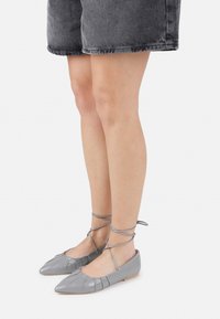 Zapatos de ballet planos y grises con puntera puntiaguda, detalle de cordones en forma de cruz alrededor del tobillo y una textura de cuero suave y brillante.