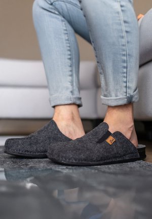 Persoon die lichtblauwe opgerolde spijkerbroek en donkergrijze instapsloffen draagt, staand op een reflecterende vloer bij een bank.