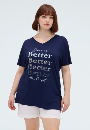 Vrouw met kort bruin haar die een donkerblauw V-hals T-shirt met de tekst "Done is Better than Perfect" draagt en witte shorts, kijkt vooruit.