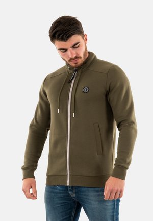 Sudadera verde oliva con cremallera y cuello alto, bolsillos frontales y un parche redondo con logo en el lado izquierdo del pecho. Tejido suave y diseño ajustado.