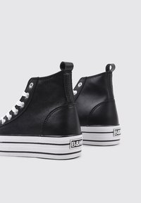 Des baskets montantes noires en cuir avec des semelles en caoutchouc blanches. Les caractéristiques comprennent des lacets blancs, des accents noir et blanc rayés, ainsi qu'un détail de logo.