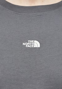 Κοντινό πλάνο γκρι υφάσματος με το λευκό κεντημένο λογότυπο "The North Face" στην περιοχή του στήθους σε μπλούζα με στρογγυλή λαιμόκοψη.