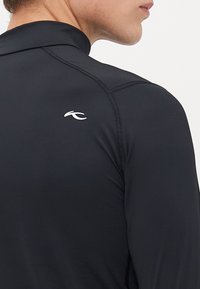 Homme portant un haut de sport ajusté à manches longues noir avec un logo blanc au dos, présentant des détails de couture visibles le long des épaules et des manches.