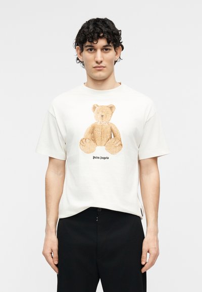 Jeune homme aux cheveux bouclés portant un t-shirt blanc avec un grand motif d'ours en peluche beige et un pantalon noir, sur un fond uni.